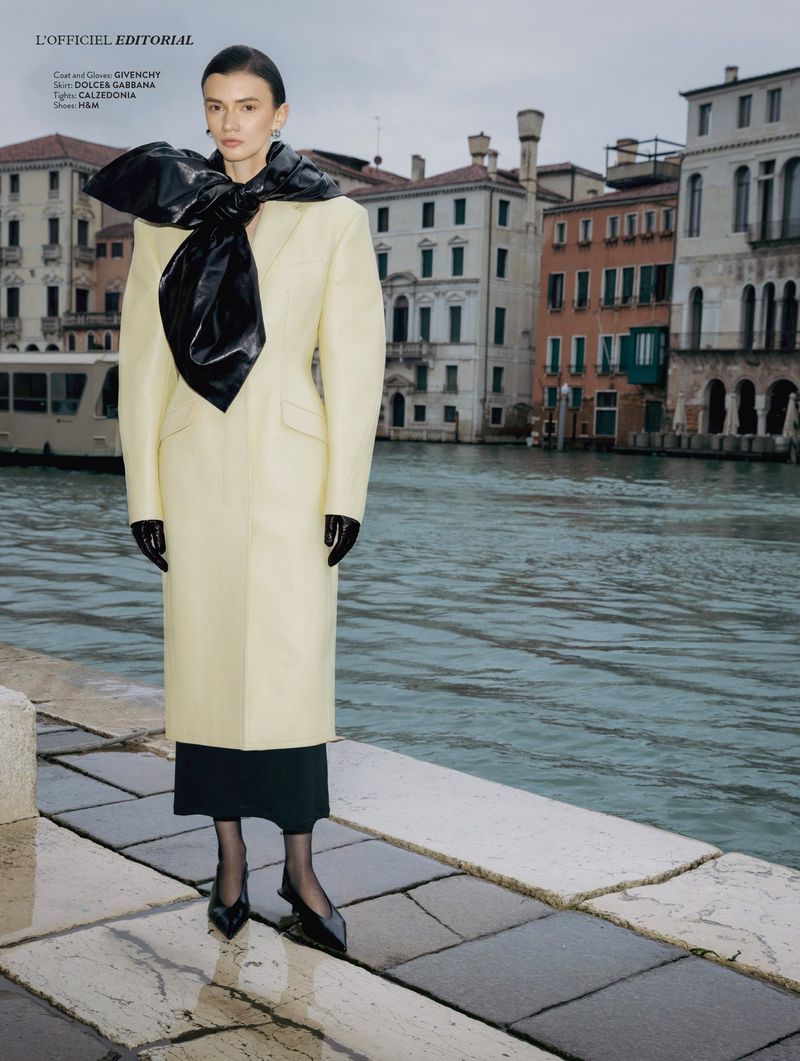 Model in a pale yellow tailored coat with an oversized black vinyl bow posing by a Venice canal on wet stone pavement.