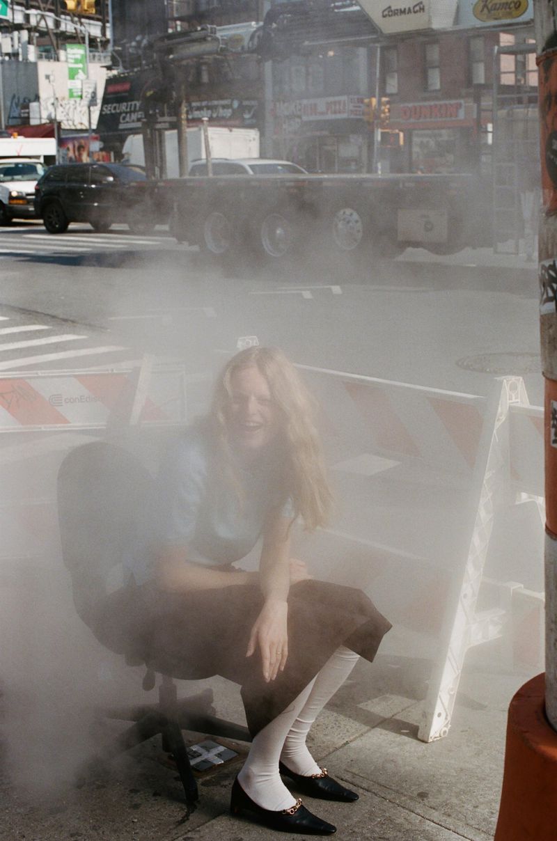 Model Hanne Gaby Odiele sitting on a wheeled office chair on a city sidewalk surrounded by dense rising steam.