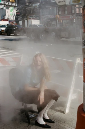 Model Hanne Gaby Odiele sitting on a wheeled office chair on a city sidewalk surrounded by dense rising steam.