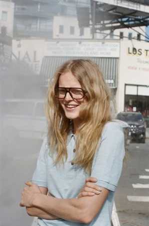 Model Hanne Gaby Odiele smiling with arms crossed wearing oversized glasses and a light blue polo shirt on a city street.