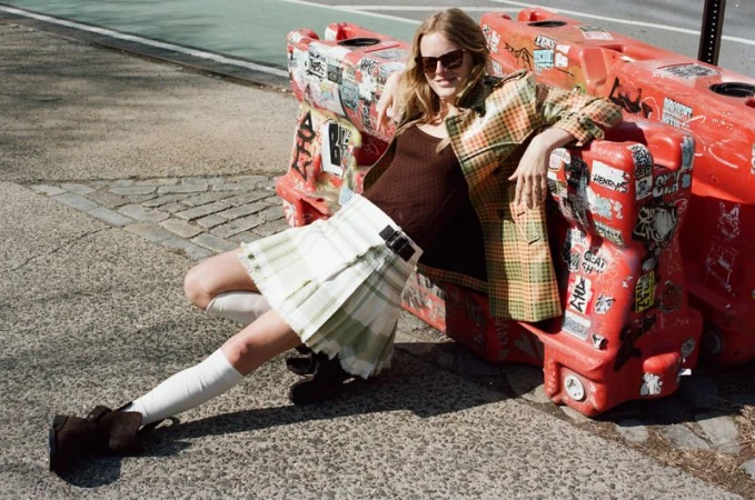 Hanne Gaby Odiele wearing a plaid coat and pleated skirt poses against red street barriers in sunglasses.