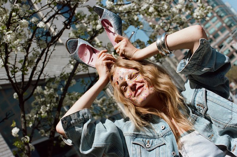 Model Hanne Gaby Odiele holds sparkling shoes above her head wearing an oversized light denim jacket outdoors near a flowering tree.
