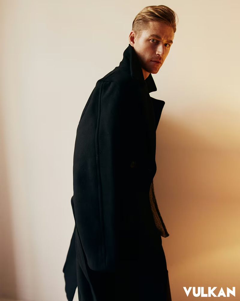Man in a black high-collar overcoat looking over his shoulder against a softly lit neutral background.