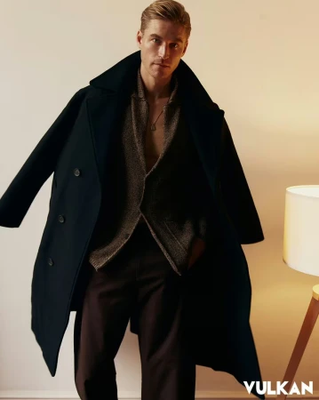 Chad John Payne wearing an open brown jacket, dark overcoat over his shoulders, and dark trousers posing beside a floor lamp.