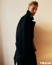 Man in a black high-collar overcoat looking over his shoulder against a softly lit neutral background.