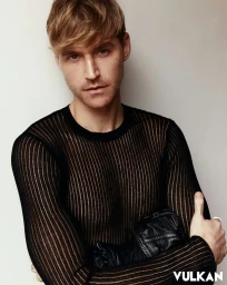 Portrait of a man with styled blonde hair in a black sheer ribbed long-sleeve top against a white studio background.