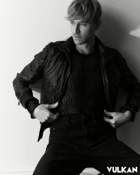 Black and white portrait of a man wearing a leather jacket and ribbed knit sweater posing against a white wall.