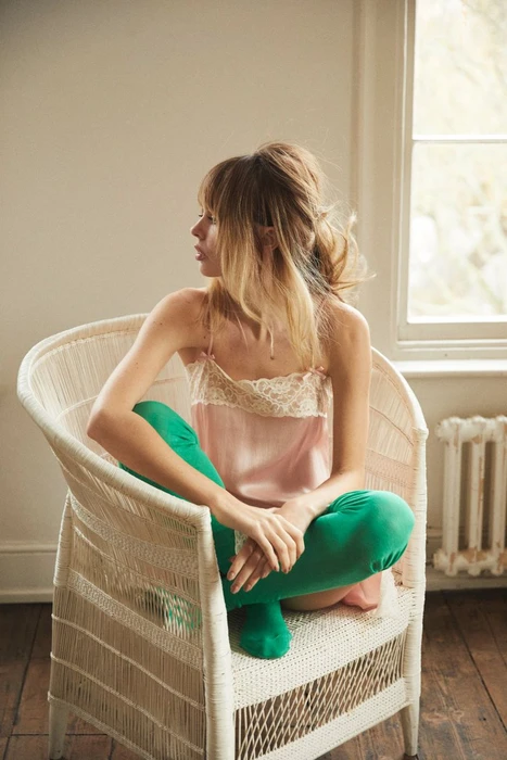 Model Izzy Hawken wearing a pink lace slip top and bright green tights seated cross-legged in a white woven wicker armchair.