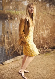 Model Izzy Hawken wearing a yellow floral shift dress and brown jacket outdoors by a reed-lined waterside.