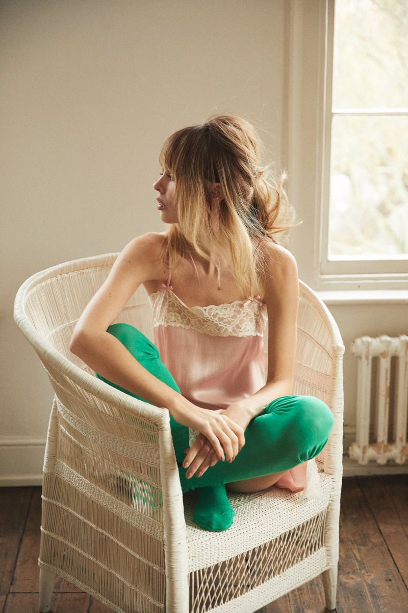 Model Izzy Hawken wearing a pink lace slip top and bright green tights seated cross-legged in a white woven wicker armchair.