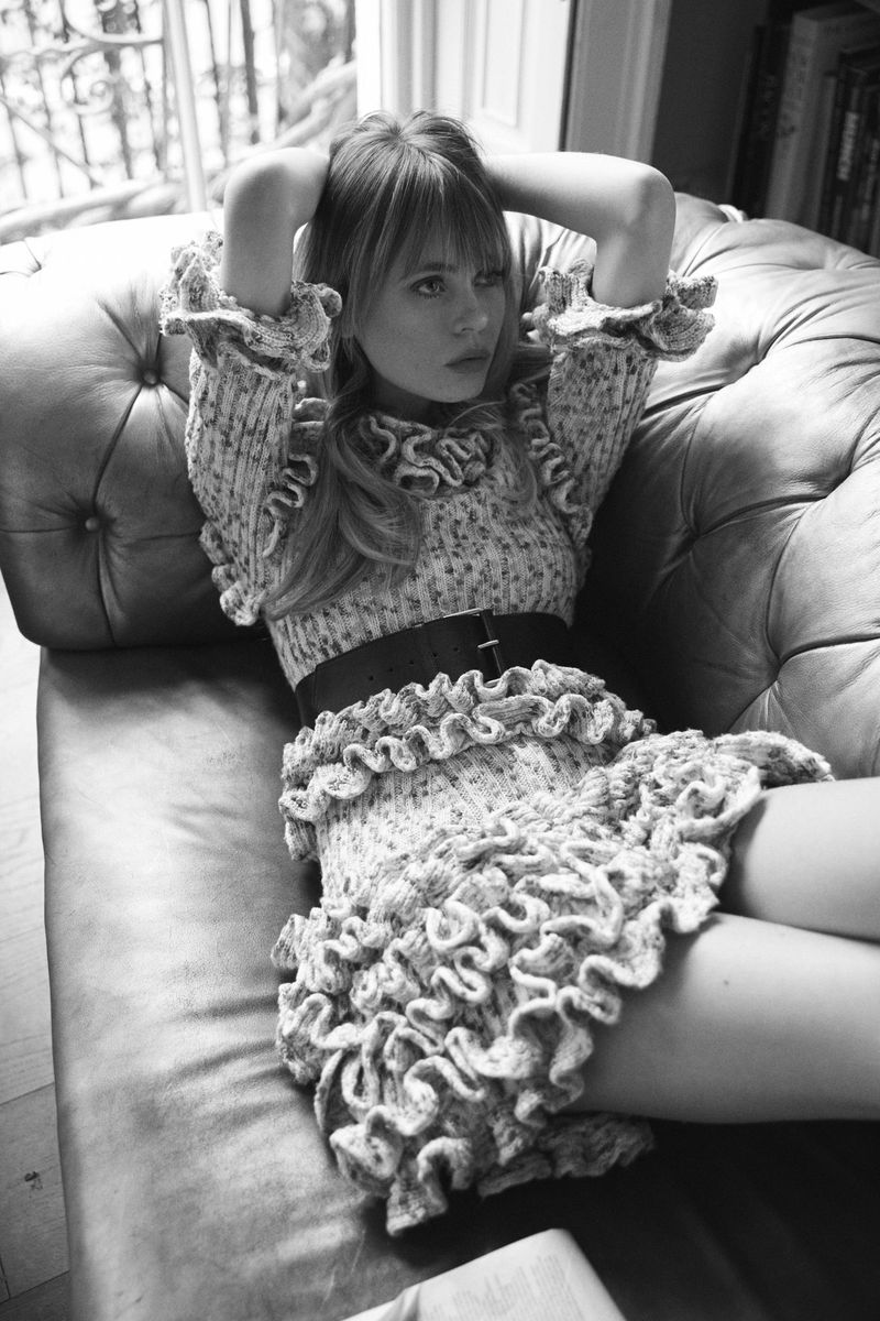 Black and white portrait of a woman wearing a ruffled dress and thick belt lounging on a tufted leather sofa.