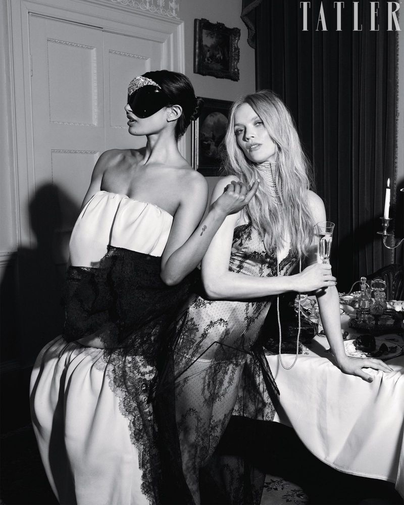 Black and white editorial photo of two models in lace eveningwear, one wearing an embellished eye mask, posing beside a dining table.