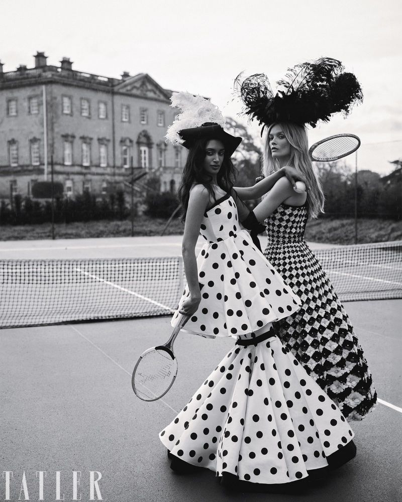 Two female models in patterned dresses and feathered headwear holding tennis rackets on a court in front of a grand house.