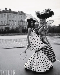 Two female models in patterned dresses and feathered headwear holding tennis rackets on a court in front of a grand house.
