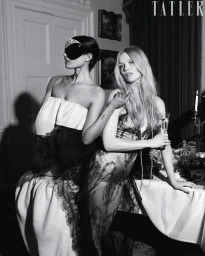 Black and white editorial photo of two models in lace eveningwear, one wearing an embellished eye mask, posing beside a dining table.