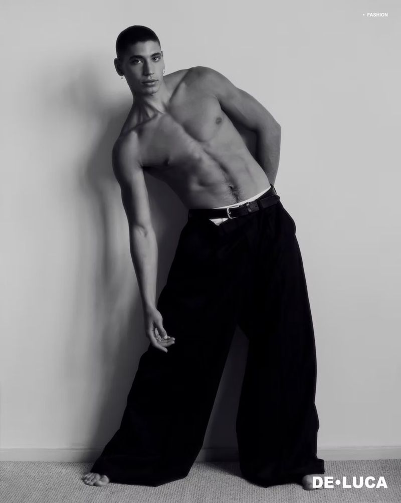 Black and white portrait of a shirtless male model with toned physique, wearing loose black pants and posing in front of a wall.