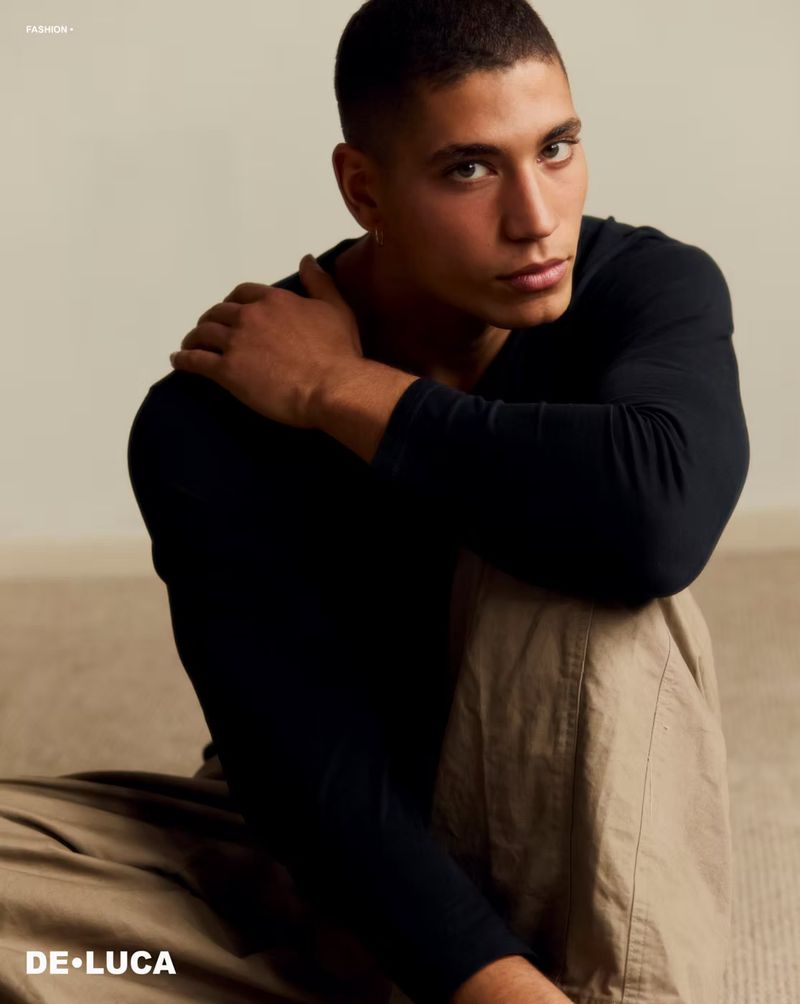 Male model wearing a long-sleeved black shirt and beige pants, seated indoors against a neutral background, looking at camera.