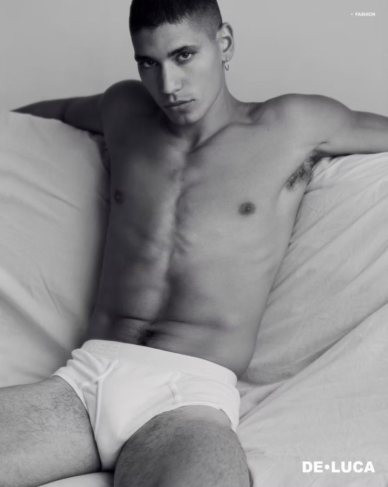 Shirtless male model Vincent Rayvon in white briefs reclining against draped white bedding in a black and white portrait.