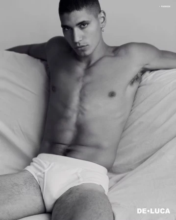 Shirtless male model Vincent Rayvon in white briefs reclining against draped white bedding in a black and white portrait.