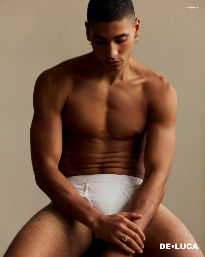 A shirtless male model poses while sitting in white briefs. The photo is lit with studio lighting against a neutral background. He wears hoop earrings.
