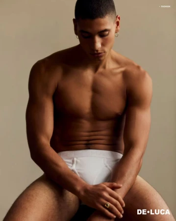 A shirtless male model poses while sitting in white briefs. The photo is lit with studio lighting against a neutral background. He wears hoop earrings.