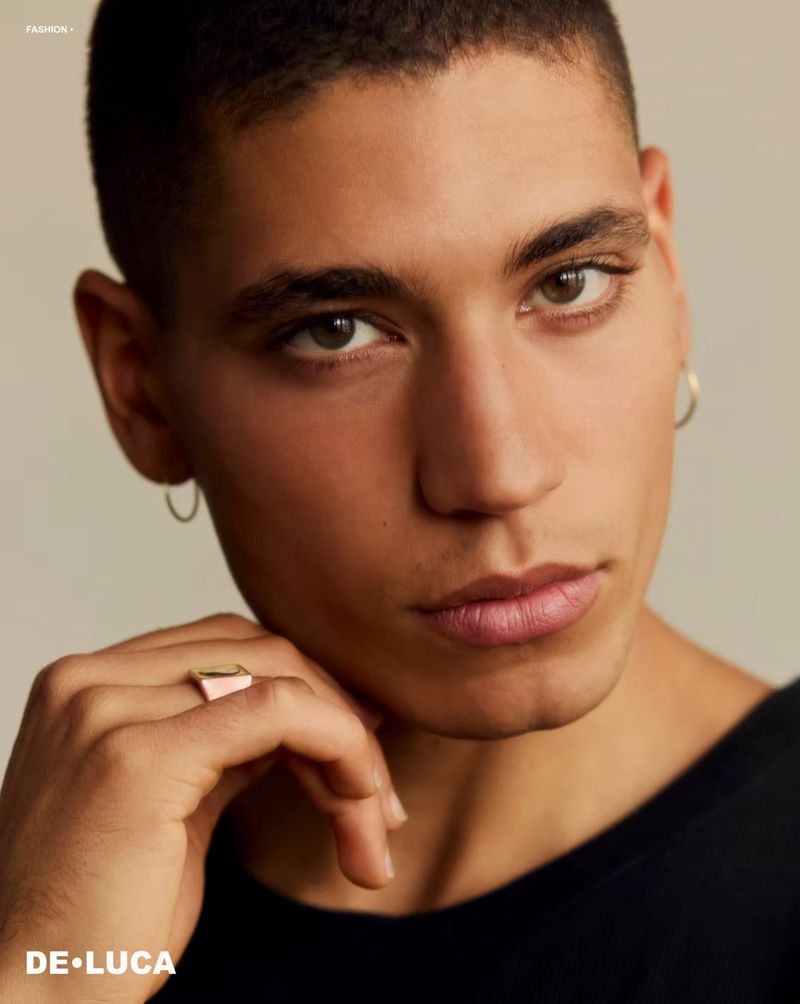 Close-up portrait of model Vincent Rayvon wearing hoop earrings, a signet ring, and a black shirt against a beige background.