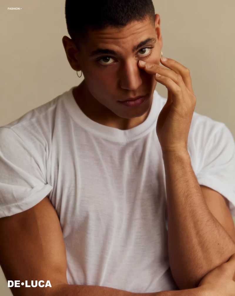 Portrait of a tanned adult male in a white t-shirt with his hand near his face and an earring, posing in front of a neutral background.