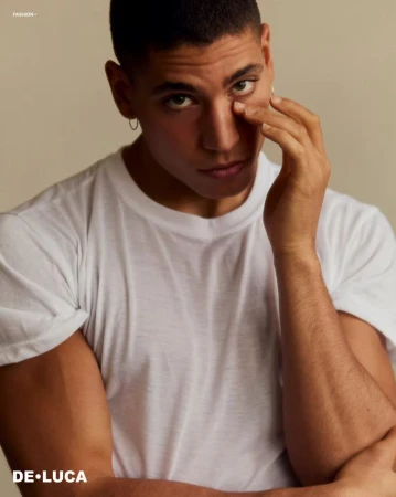 Portrait of a tanned adult male in a white t-shirt with his hand near his face and an earring, posing in front of a neutral background.