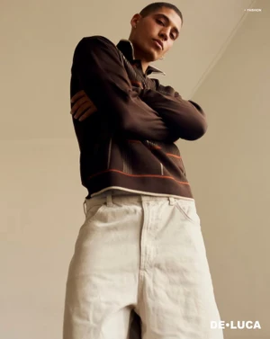 Low-angle shot of a male model with crossed arms wearing a patterned dark top and white trousers against a neutral wall.