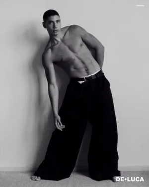 Shirtless male model posing against a plain wall wearing wide-cut dark trousers and a belt.