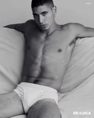 Shirtless male model Vincent Rayvon in white briefs reclining against draped white bedding in a black and white portrait.