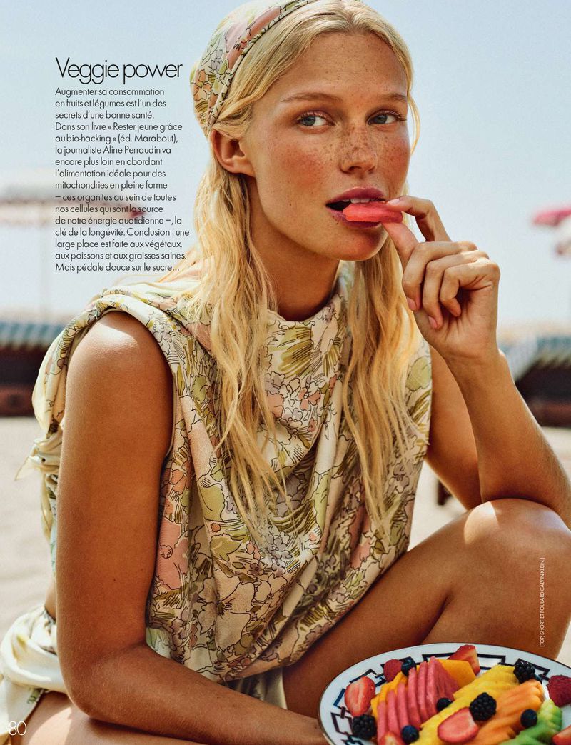 Blonde woman with freckles in a floral top and headscarf eating fruit on a sunny beach.