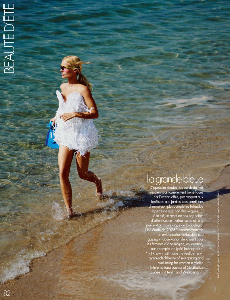 Model in a white feathered strapless minidress running along the ocean shoreline on a sandy beach holding blue swim fins.