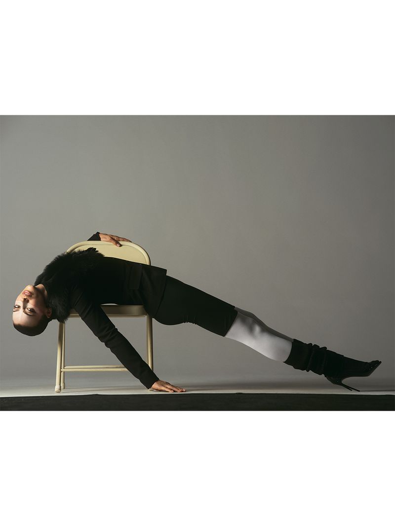 Irina Shayk arching backward over a beige chair in a black outfit, white tights, and black stiletto boots against a studio backdrop.
