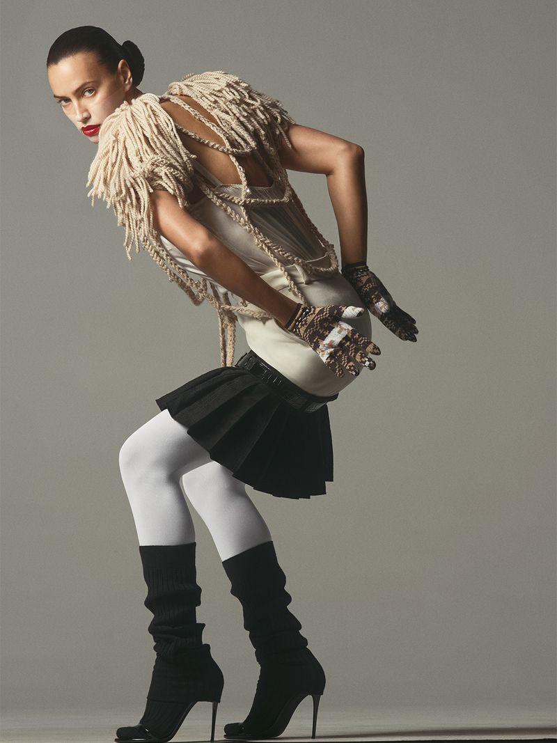 Model Irina Shayk posing in a textured cream fringe top, black pleated miniskirt, white tights, and black high heels in studio.