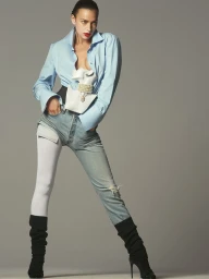 Irina Shayk posing in a light blue button-down shirt, deconstructed denim jeans, and black stiletto boots against a grey studio backdrop.