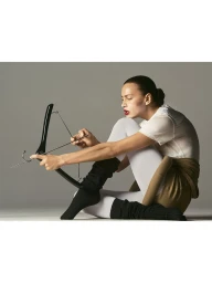 Irina Shayk sitting in white tights and black leg warmers holding a black clothes hanger in front of a neutral grey backdrop.