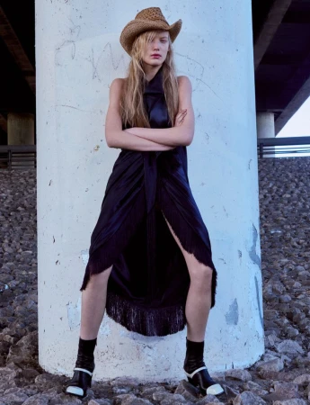 Model Alina Miller in a black fringe dress and straw cowboy hat leaning against a concrete pillar under a bridge.