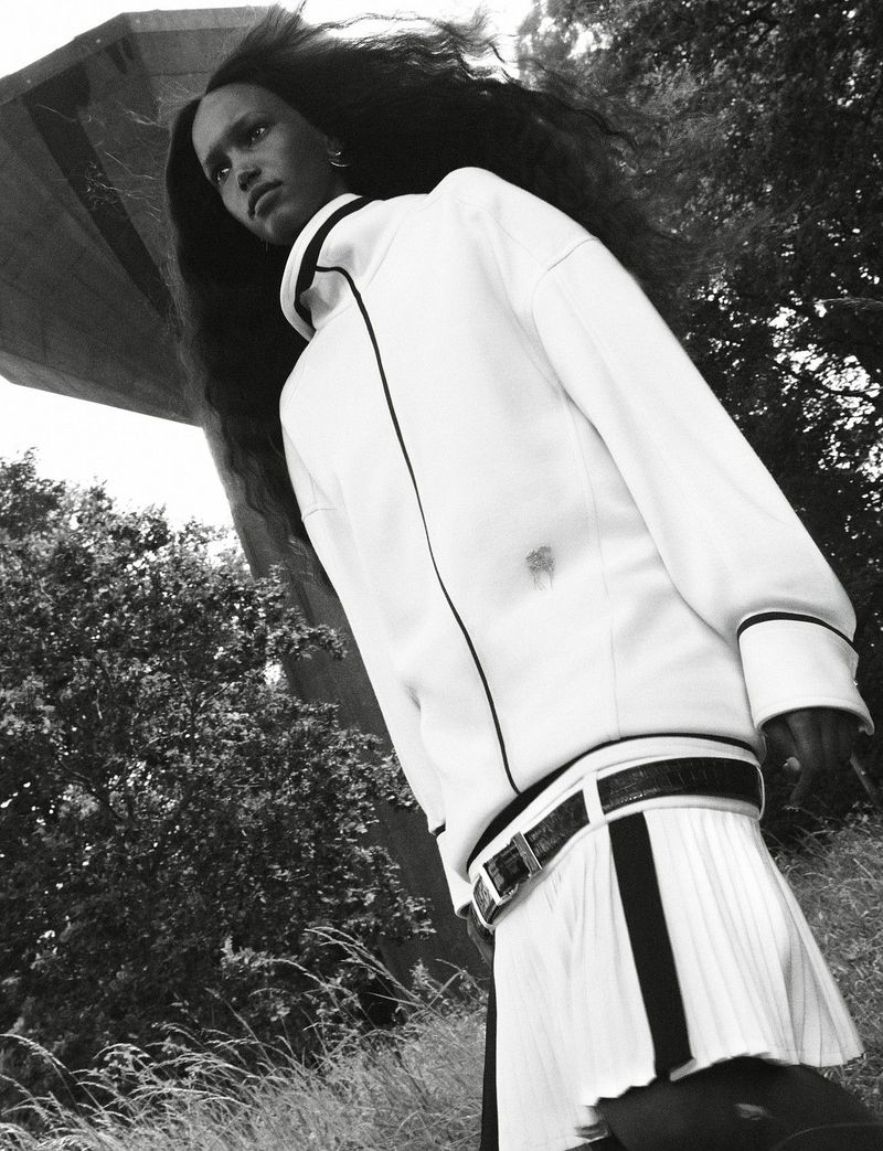 Black and white low-angle photograph of a woman in a white sporty zip jacket and pleated skirt standing outdoors near trees.