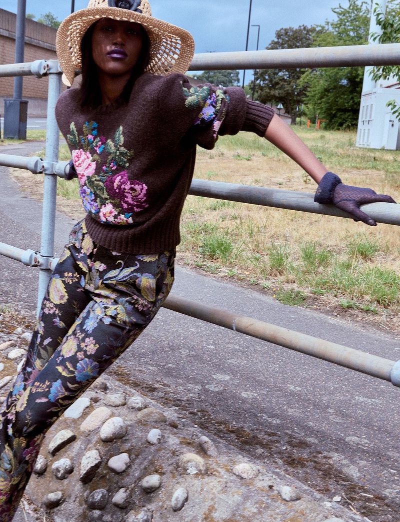 Model in a brown floral sweater and straw hat leaning against a metal railing beside an outdoor road.