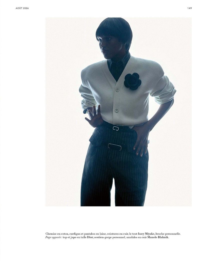 Model posing in a white cardigan with a black flower brooch and dark pinstripe trousers against a bright white background.