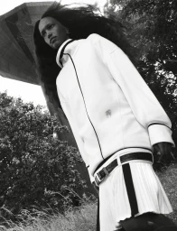 Black and white low-angle photograph of a woman in a white sporty zip jacket and pleated skirt standing outdoors near trees.