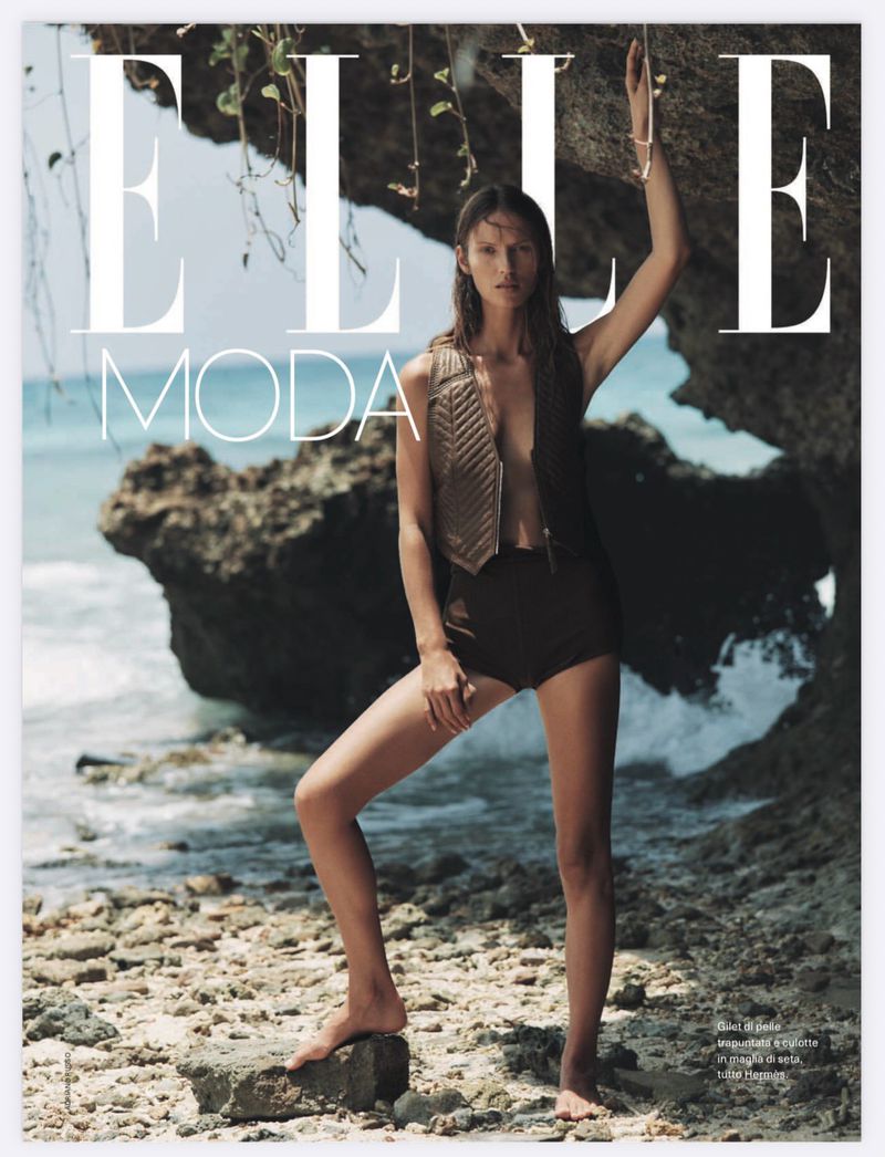 Model standing on rocky shore wearing dark briefs and a sleeveless quilted vest with ocean background