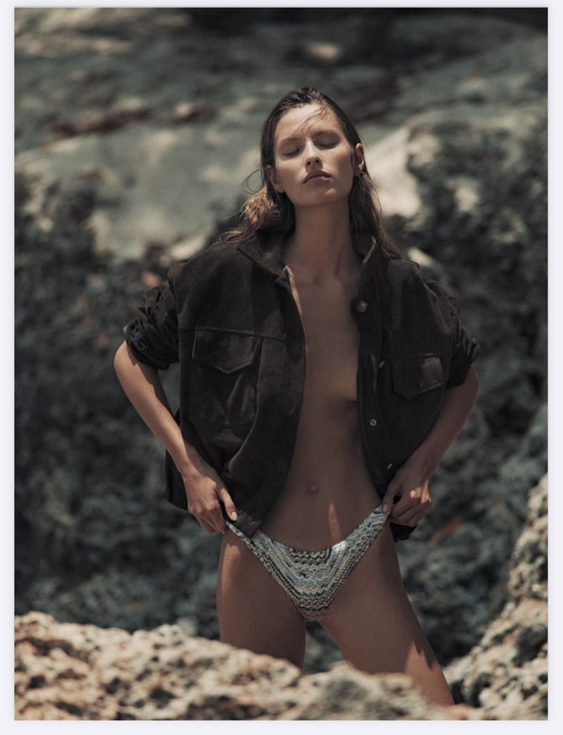 Model standing among rocky terrain wearing an open dark jacket and patterned bikini bottoms, eyes closed.