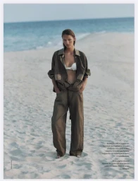 Model Lea Seelenmeyer stands barefoot on a sandy beach wearing a loose brown jacket, wide-leg trousers, and a white bikini top.
