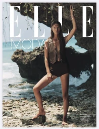 Model standing on rocky shore wearing dark briefs and a sleeveless quilted vest with ocean background