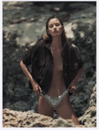 Model standing among rocky terrain wearing an open dark jacket and patterned bikini bottoms, eyes closed.