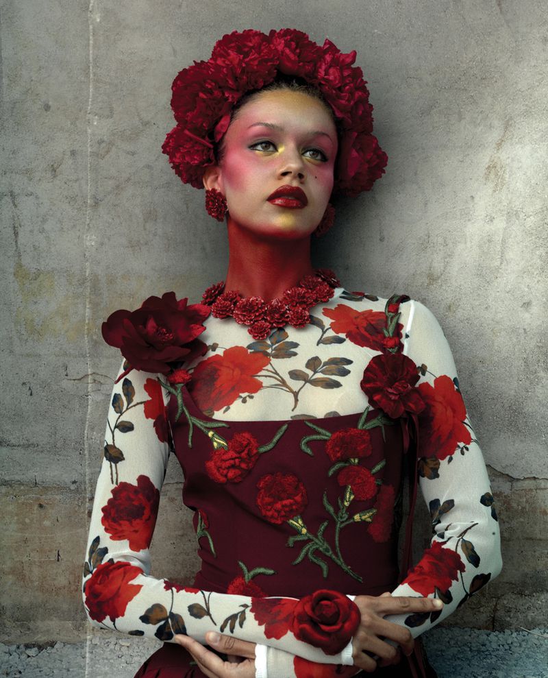 Rachel Zegler wearing a red floral embroidered top, floral headdress, and crimson neck makeup against a textured concrete wall.