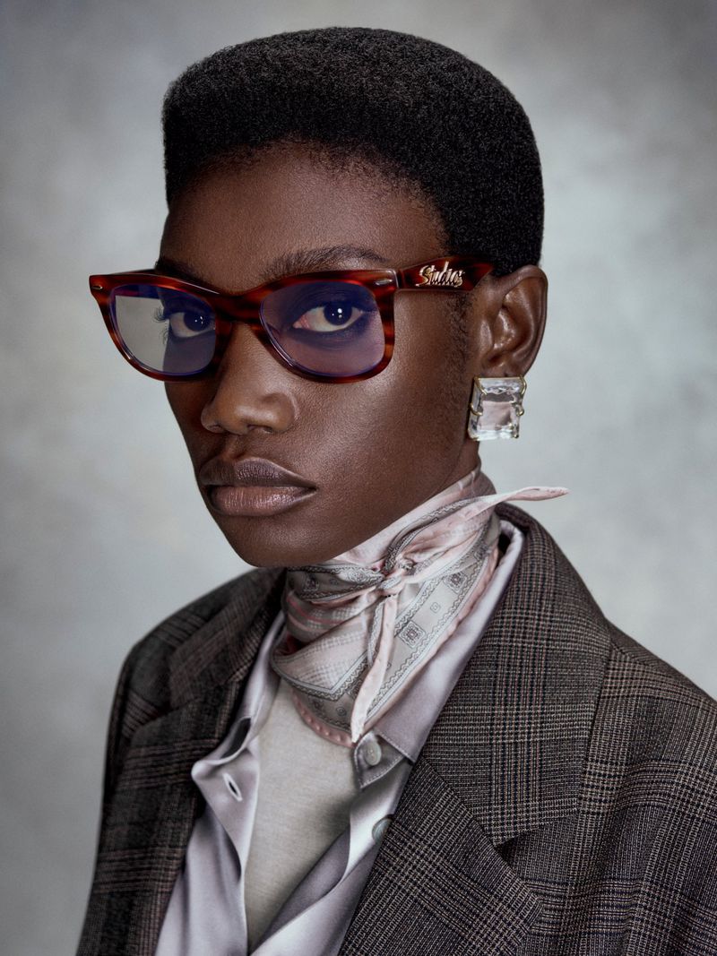 Kris Krystal wears tortoiseshell glasses, a silk neck scarf, and a plaid blazer against a mottled grey studio backdrop.
