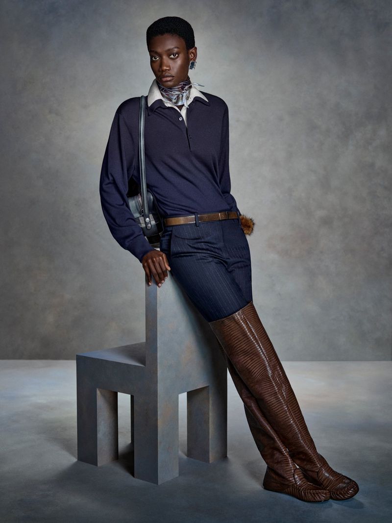 Kris Krystal wears a navy polo, striped shorts, and brown croc-embossed thigh-high boots while leaning on a geometric grey chair.